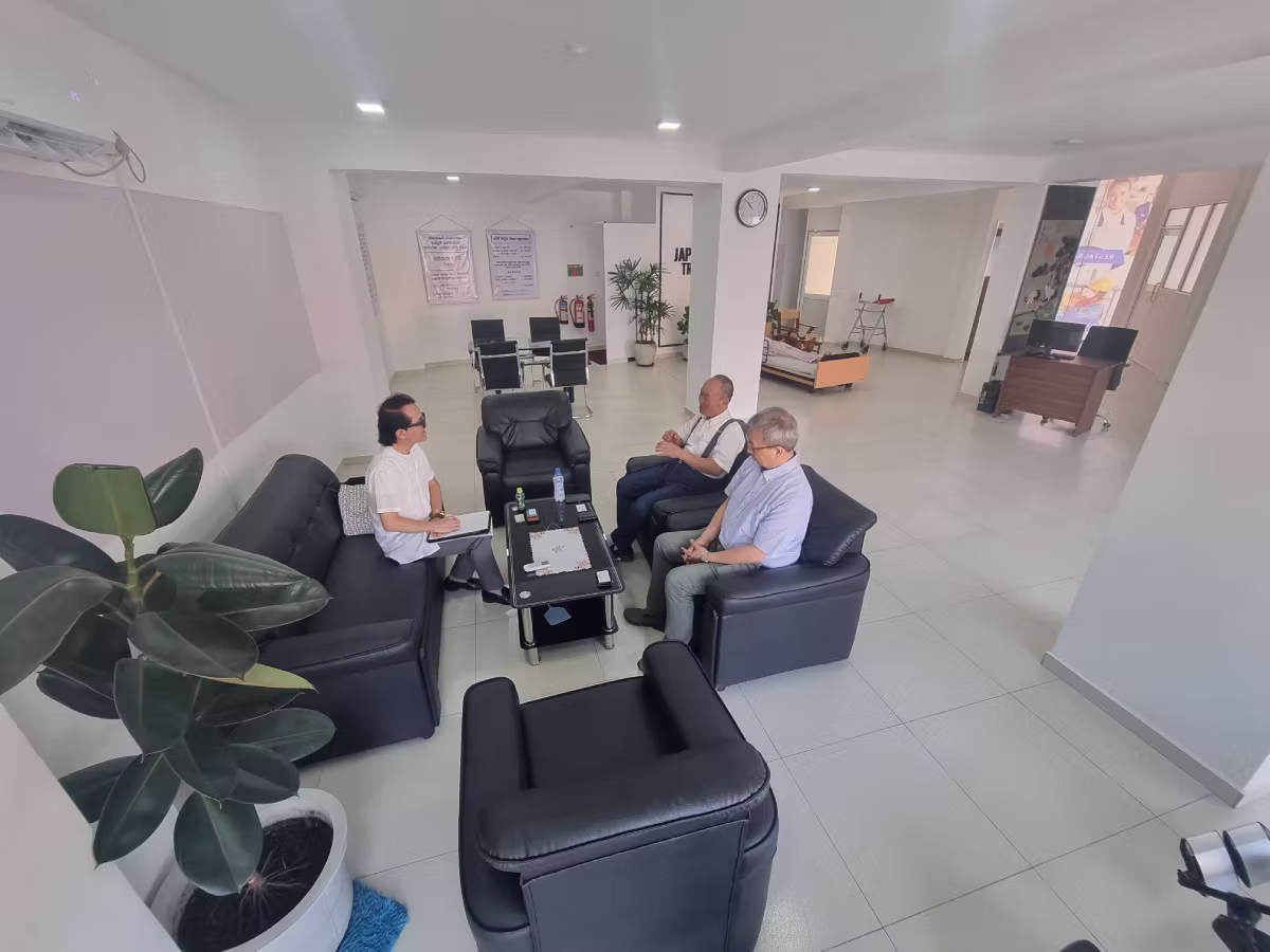 Three men in business casual attire sit on black leather seating in a bright, modern office waiting area with white walls and tiled floors.